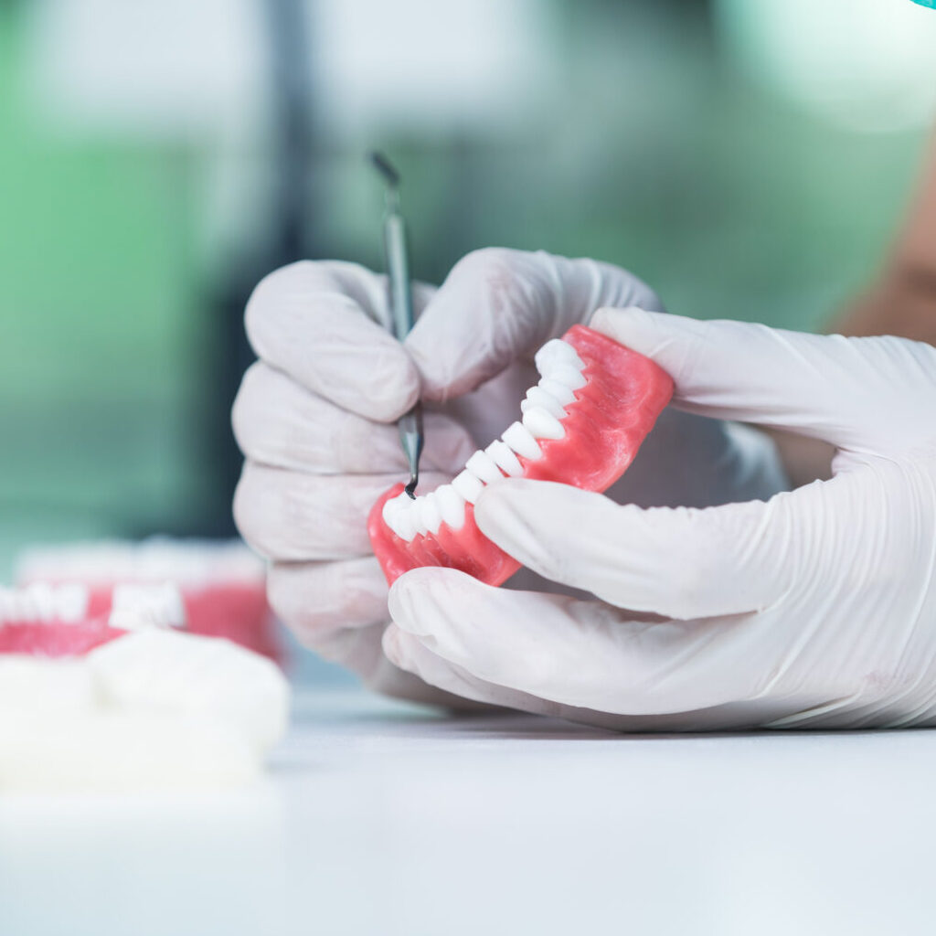 Dental Prosthesis, Dentures, Prosthetics Work. Prosthetics Hands While Working On The Denture, False Teeth, A Study And A Table With Dental Tools. Dental Prosthesis, Dentures, Prosthetics Work. Prosthetics Hands While Working On The Denture, False Teeth, A Study And A Table With Dental Tools.